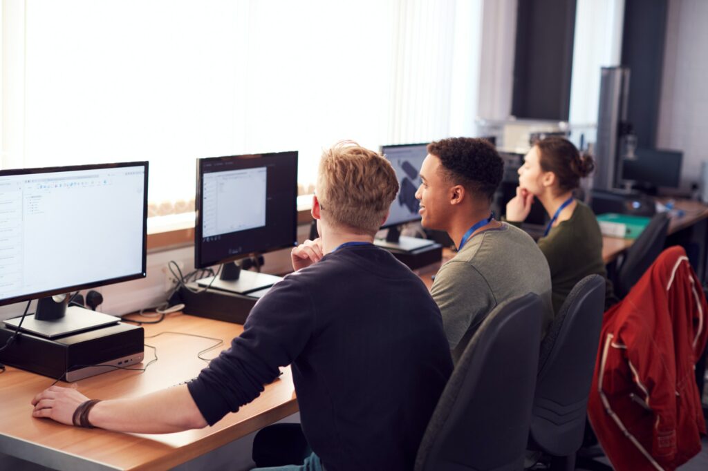 Group Of College Students Studying Computer Design Sitting At Line Of Monitors In Classroom